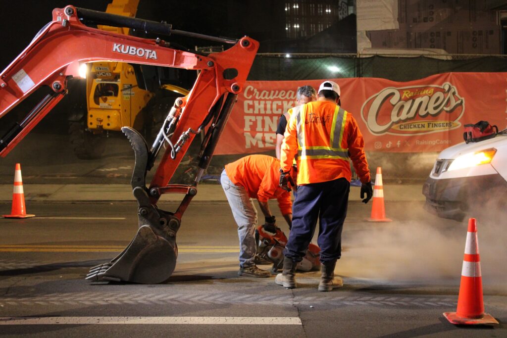 Trinity crew saw-cutting asphalt beside an excavator at Raising Cane's on Broadway