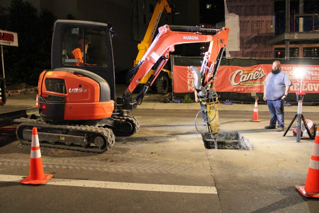 Mini excavator working inside a coned lane closure on Broadway at night